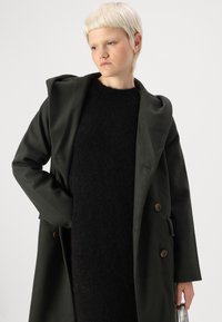 Mørkegrønn frakk med dobbelt knappdesign, to brune knapper og hette. Kombinert med en svart strukturert genser under.