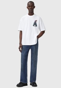 Hvid bomulds t-shirt med et grafisk print, der viser en figur og tekst. Parret med slidte blå denim jeans og sort fodtøj.