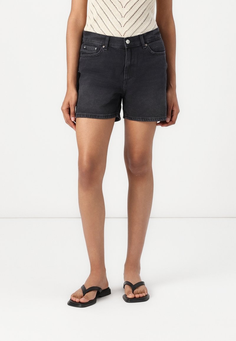 Personne portant un short en jean noir, un haut sans manches en maille crème et des sandales plates noires, debout devant un fond blanc.