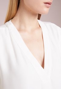 Närbild av en kvinnas hals och bröst som bär en vit V-ringad blus med långt rakt blont hår synligt.