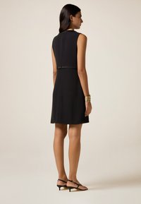Schwarzes ärmelloses Kleid mit tailliertem Oberteil und schwingendem Rock, mit einem Taillendetail. Das Model trägt schwarze Riemchenheels.