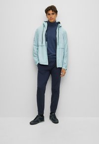 Gaiši zila neilona jakas ar rāvējslēdzēju un kapuci, tumši zils turtlenecks, tumši zili bikses un tumšas sporta apavus. Minimālisma dizains; nav rakstu.