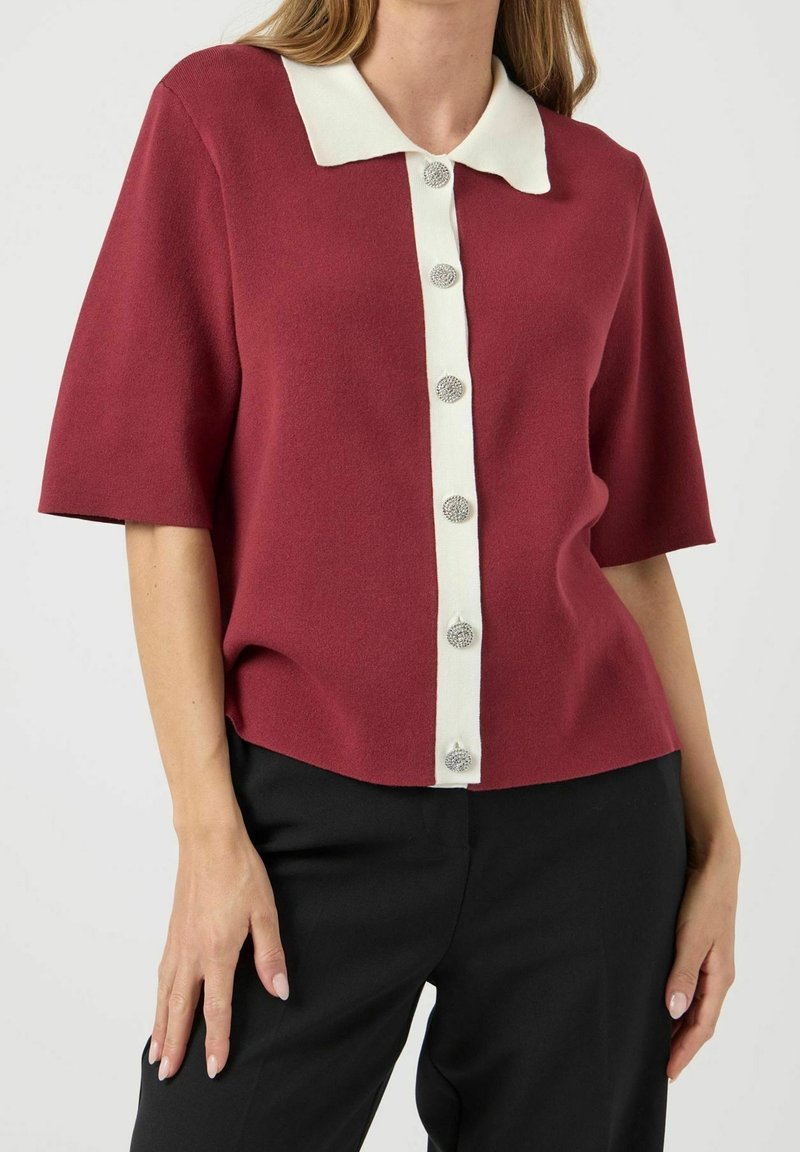 Vrouw draagt een bordeauxrode blouse met korte mouwen, witte kraag en knoopsluiting, gecombineerd met een zwarte broek, staand tegen een witte achtergrond.