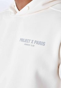 Witte hoodie van zacht materiaal, met een kangaroo pocket en een trekkoord capuchon. Tekst "PROJECT X PARIS MEMBERS' CLUB" in grijs op de voorkant.