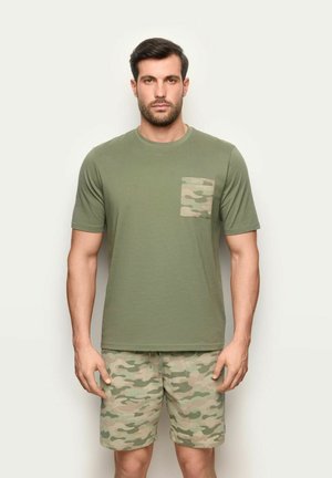 Mann trägt ein olivgrünes T-Shirt mit Tarnflecktasche und passende Tarnfleck-Shorts, steht vor einem einfarbigen hellen Hintergrund.