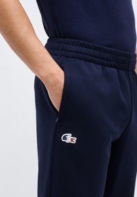 Ενήλικας που φοράει σκούρο μπλε φόρμα με ελαστική μέση και χέρι στην τσέπη, με μικρό τρίχρωμο λογότυπο κροκόδειλου στον μηρό.