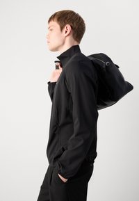 Čierna ľahká bunda s vysokým golierom, elastickými manžetami a zipsovými bočnými vreckami, v kombinácii s elegantným čiernym batohom.