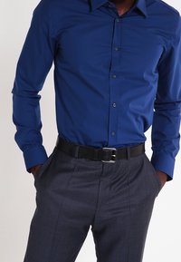 Herren-Langarmhemd in Blau, tailliertes Design, glatte Textur, durchgehende Knopfleiste, kombiniert mit grauen, gemusterten Hosen und einem schwarzen Gürtel.