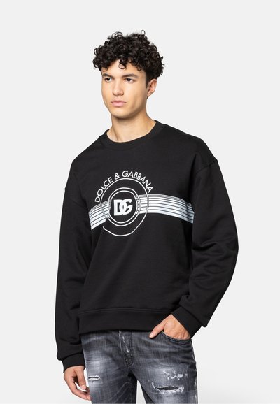 Zwarte sweatshirt met een ronde hals, geribbelde manchetten en een borstlogo met witte accenten en strepen. Draagt met versleten grijze jeans.