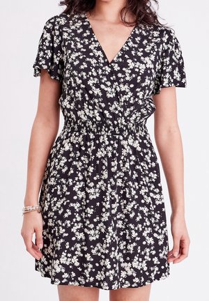 Femme portant une robe noire à manches courtes avec imprimé floral blanc et taille élastique, bras détendus le long du corps avec des bracelets au poignet gauche.