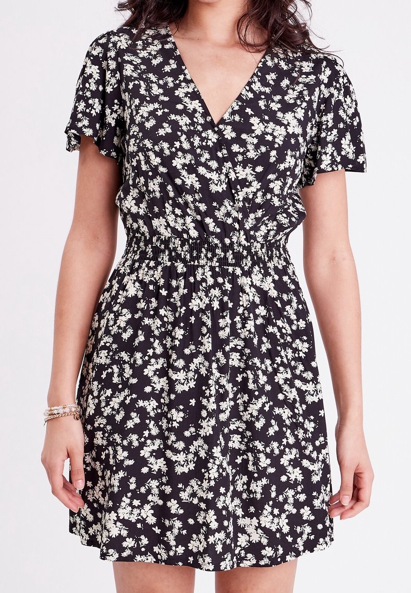 Femme portant une robe noire à manches courtes avec imprimé floral blanc et taille élastique, bras détendus le long du corps avec des bracelets au poignet gauche.