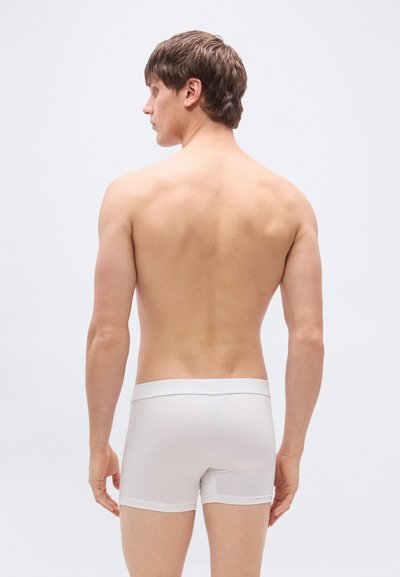 Boxers blancs en coton avec une ceinture douce, un design ajusté et une texture lisse. Le modèle est de dos, cheveux courts, sur un fond gris.