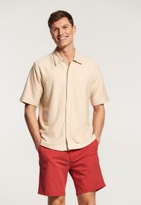 Camisa corta de manga corta color beige claro con tejido texturizado, cierre de botones y cuello puntiagudo, combinada con pantalones cortos rojos.