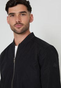 Schwarze Bomberjacke mit Reißverschluss vorne, geripptem Kragen und strukturiertem Stoff; verfügt über ein rundes Logo-Patch am linken Ärmel.