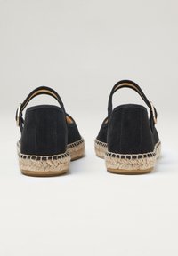 Černé semišové espadrilky s jutanovou podrážkou, které mají zadní řemínek a boční přezku. Měkká textura s lehkým, neformálním designem.