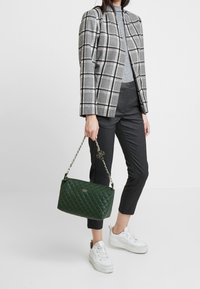 Sac en bandoulière matelassé vert avec une chaîne, associé à un blazer à carreaux gris et un haut gris, accompagné de pantalons rayés et de baskets blanches.