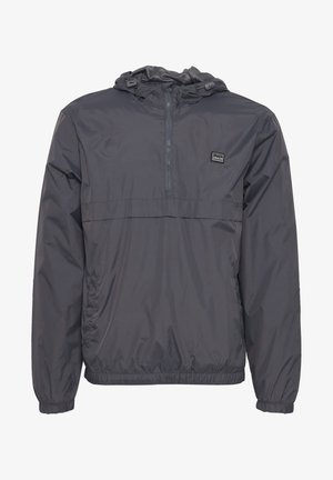 Giacca leggera grigia con cappuccio e chiusura a mezza zip, dotata di polsini elasticizzati. Presenta un piccolo patch con logo sul petto.