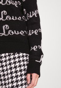 Černý pletený svetr se vzorem "Láska" v bílé barvě, s žebrovanými manžetami. V kombinaci s sukní s kostkovaným vzorem v černé a bílé barvě.