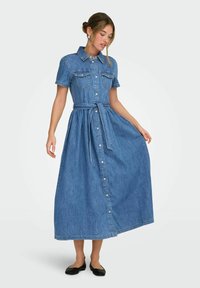 Femme portant une robe midi en denim bleu avec des boutons sur le devant, à manches courtes et une ceinture à la taille, associée à des ballerines noires, debout devant un fond blanc.