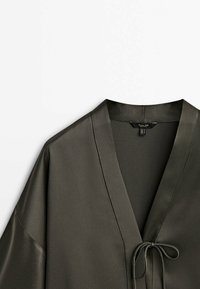 Blouse en satin vert olive foncé avec col en V et fermeture fine à nouer à l’avant, présentée sur fond blanc.