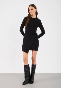 Čierne dlhé šaty s dlhými rukávmi a zavinovacím dizajnom, vybavené opaskom na viazanie v páse a asymetrickým lemom. Kombinované s čiernymi čižmami po kolená. Hladká textúra.