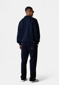 Mann in einem marineblauen Trainingsanzug und weißen Turnschuhen steht mit dem Rücken zur Kamera vor einem weißen Hintergrund, mit Händen in den Taschen.