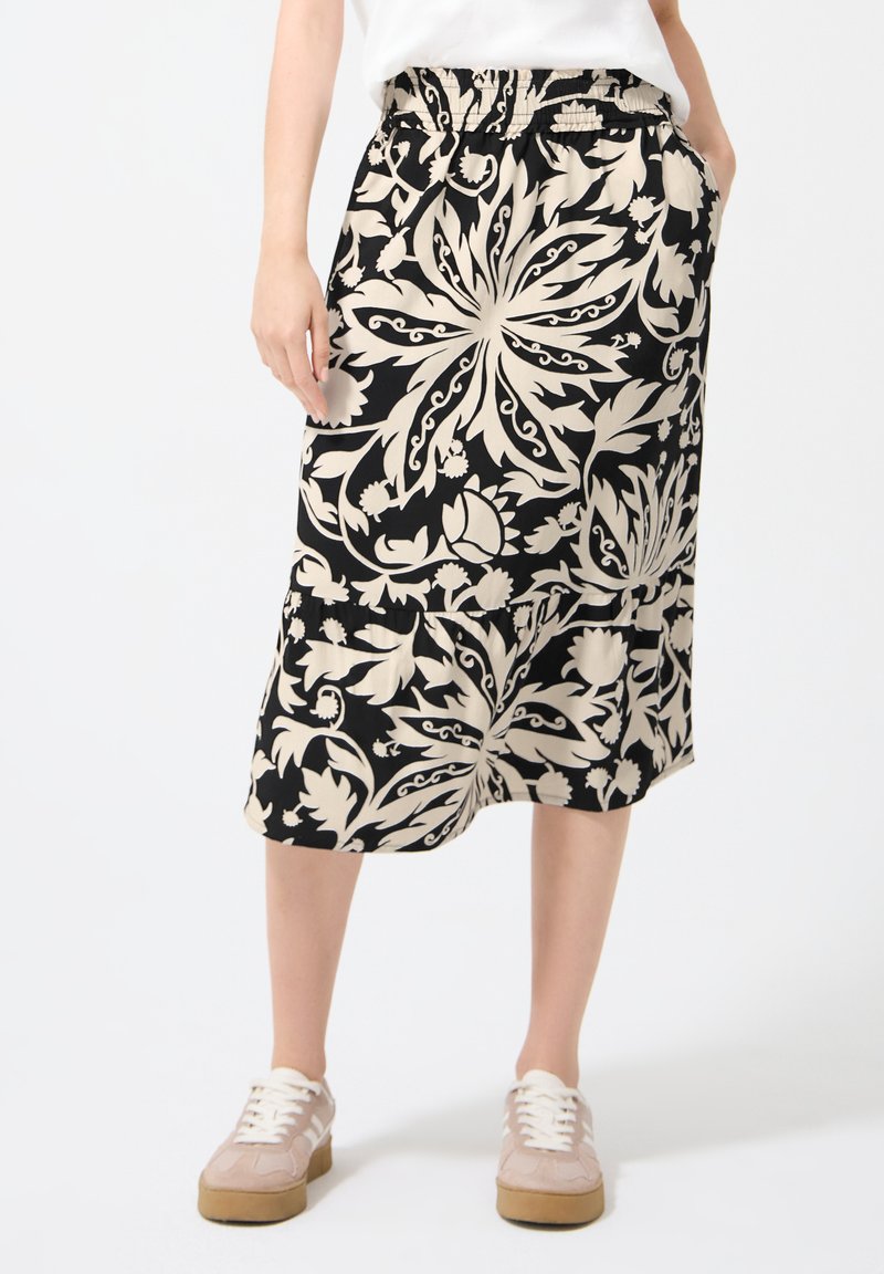 Femme portant une jupe midi noire avec un motif floral beige, taille élastique, poches latérales, haut blanc et baskets beiges.