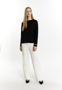 Schwarzer Pullover mit weißem Rand, kombiniert mit weißen weit geschnittenen Hosen und schwarzen spitzen Pumps. Das Model steht vor einem neutralen Hintergrund.