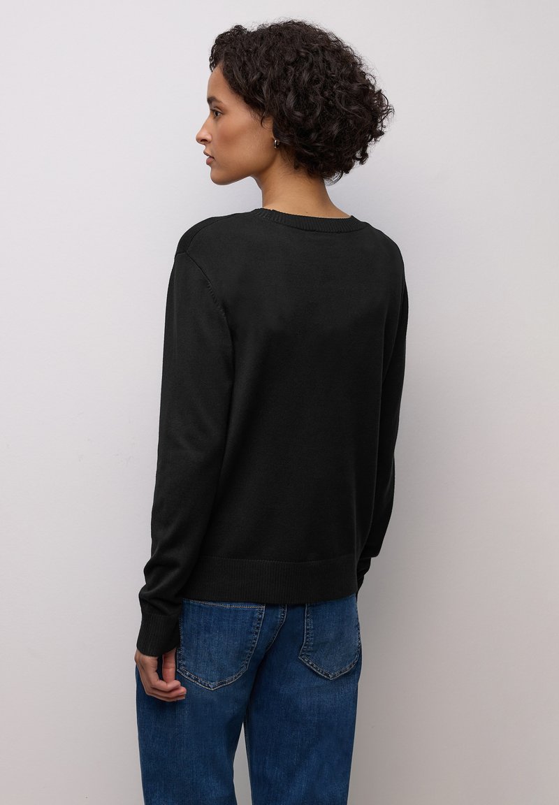 Pull noir en tricot avec manches longues, col et ourlet côtelés. Porté avec un jean denim bleu, mettant en valeur une coupe décontractée et une texture douce.