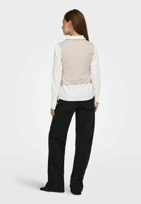 Un gilet court beige clair sur une chemise blanche à col, associé à un pantalon noir à jambe large. Texture lisse sans motifs ni accents visibles.