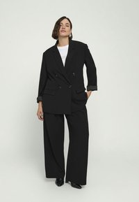 Černý dvouřadý blazer se širokými klopami, spárovaný s volnými černými kalhotami. Rukávy s manžetami mají kontrastní pruh. Pod ním je jednoduché bílé tričko.