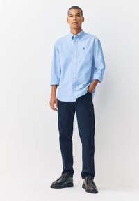 Lichtblauw overhemd met knopen en een kraag, gecombineerd met donkerblauwe chinos en zwarte veterschoenen. Gladde stof; losse pasvorm.