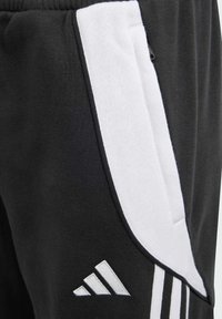 Pantalons de survêtement noirs avec une bande latérale blanche, une poche zippée et le logo Adidas en blanc sur la jambe inférieure. Texture de tissu douce.