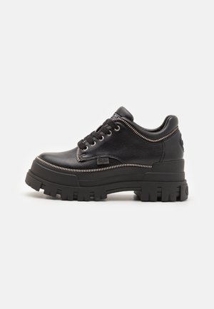 Sort lavtopet lædersneaker med kraftig rillede sål, snørelukning foran og dekorativ lynlåsdetalje langs kanterne, synligt sideskilt med mærke.