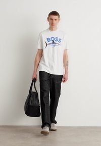 Camiseta gráfica blanca con "BOSS" en azul y un diseño de pez, combinada con jeans oscuros. El modelo lleva un bolso negro y calza zapatillas color beige.