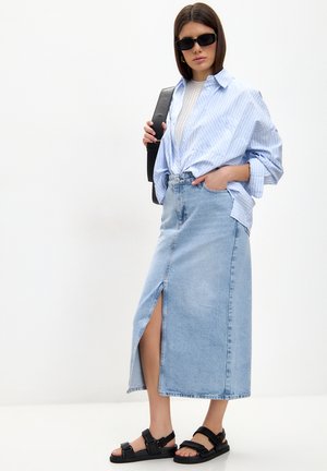 Gonna in denim azzurro chiaro con spacco frontale, abbinata a una camicia larga a righe blu e bianche, sandali neri e occhiali da sole.