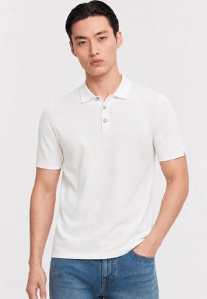 Jeune homme aux cheveux noirs coiffés en arrière, portant un polo blanc à manches courtes et un jean bleu, debout devant un fond uni.