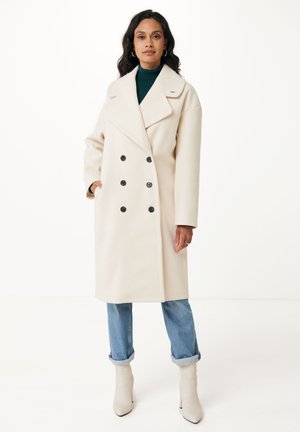 Dubbelzijdig, crèmekleurige jas met grote zwarte knopen, brede revers en zijzakken, gecombineerd met lichtblauwe jeans met manchet en witte enkellaarsjes.