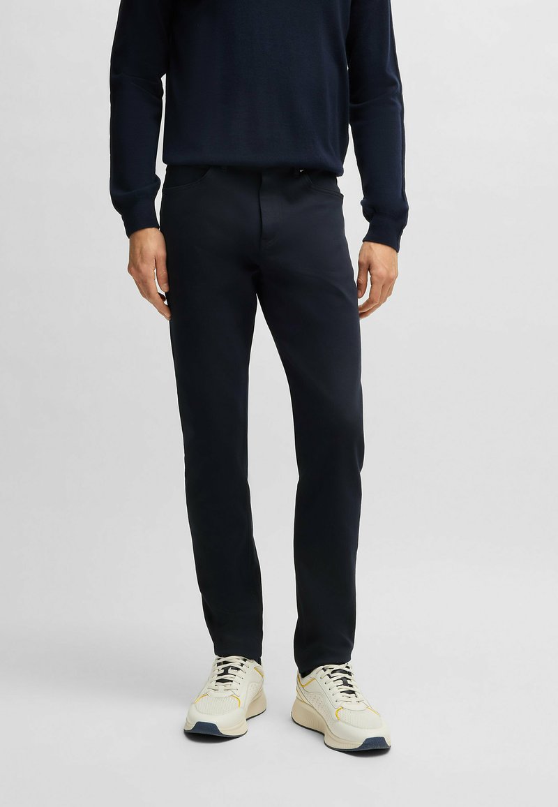 Pantalons navy en tissu lisse, coupe ajustée, avec poches avant et fermeture à bouton, associés à des baskets de couleur claire.