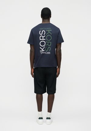 Άνδρας που φοράει μπλε σκούρο μπλουζάκι με μεγάλο κάθετο κείμενο "MICHAEL KORS" στην πλάτη, μαύρο σορτς, λευκές κάλτσες και λευκά αθλητικά παπούτσια.