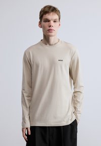 Hosszú ujjú bézs színű póló puha anyagból, kerek nyakkivágással és egy kis fekete "BOSS" logóval a bal mellrészen.