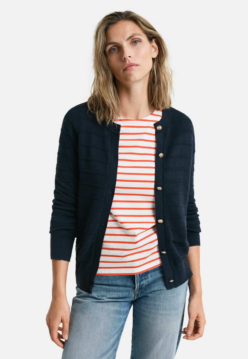 Marineblaue Strickjacke mit geripptem Muster, goldenen Knöpfen, kombiniert mit einem weiß-rot gestreiften T-Shirt, getragen zu hellblauen Jeans.