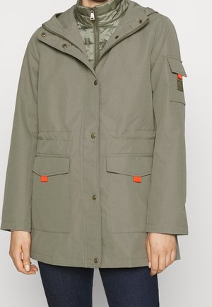 Frau trägt olivgrüne Kapuzenjacke mit vorderen Druckknöpfen, Reißverschlussfutter, Klappentaschen mit orangen Laschen und Jeans.