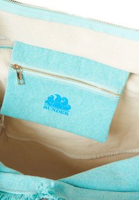 El interior de una bolsa de lienzo turquesa presenta un pequeño bolsillo con cremallera a juego con un logo azul. El forro es de color crema y suave.