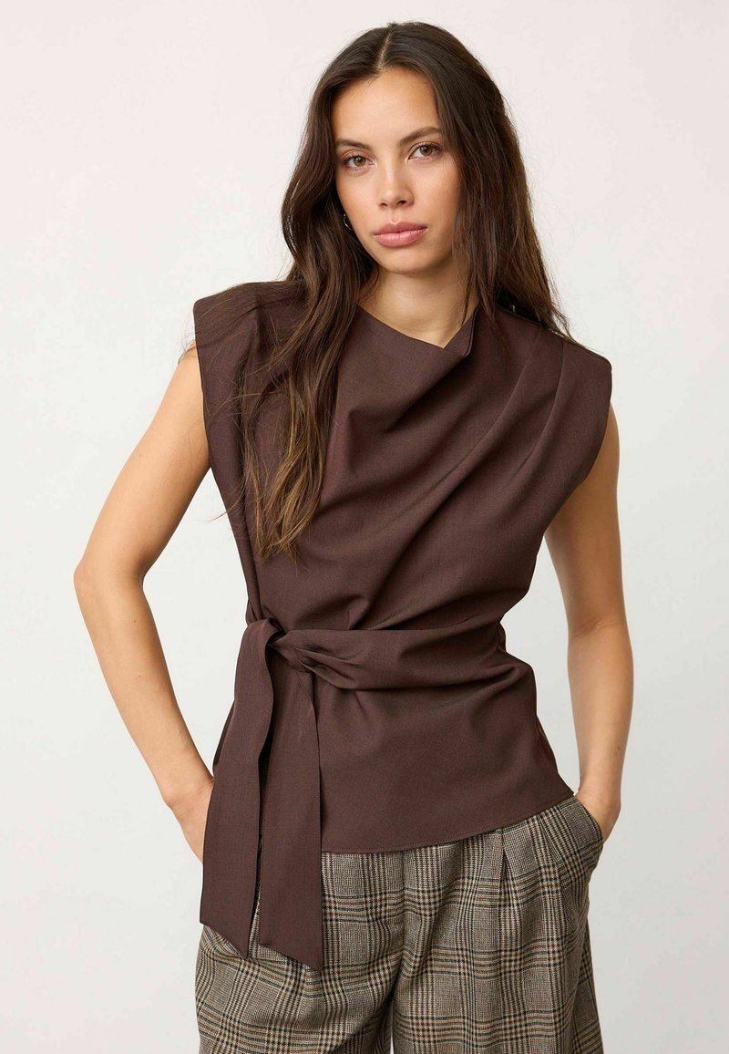 Femme aux longs cheveux bruns porte un haut marron foncé sans manches avec un lien à la taille et un pantalon à carreaux, debout avec une main dans la poche.