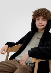 Zwarte, fuzzy zip-up jas met een capuchon, gedragen over een lichtgroene t-shirt met de tekst "NO MATTER," gecombineerd met een bruine broek.