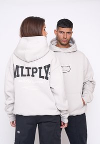 Lys grå oversized hoodie med sort tekst på ryggen, der viser et logo. Blødt stof, hætte med snoretræk og kængurulomme.