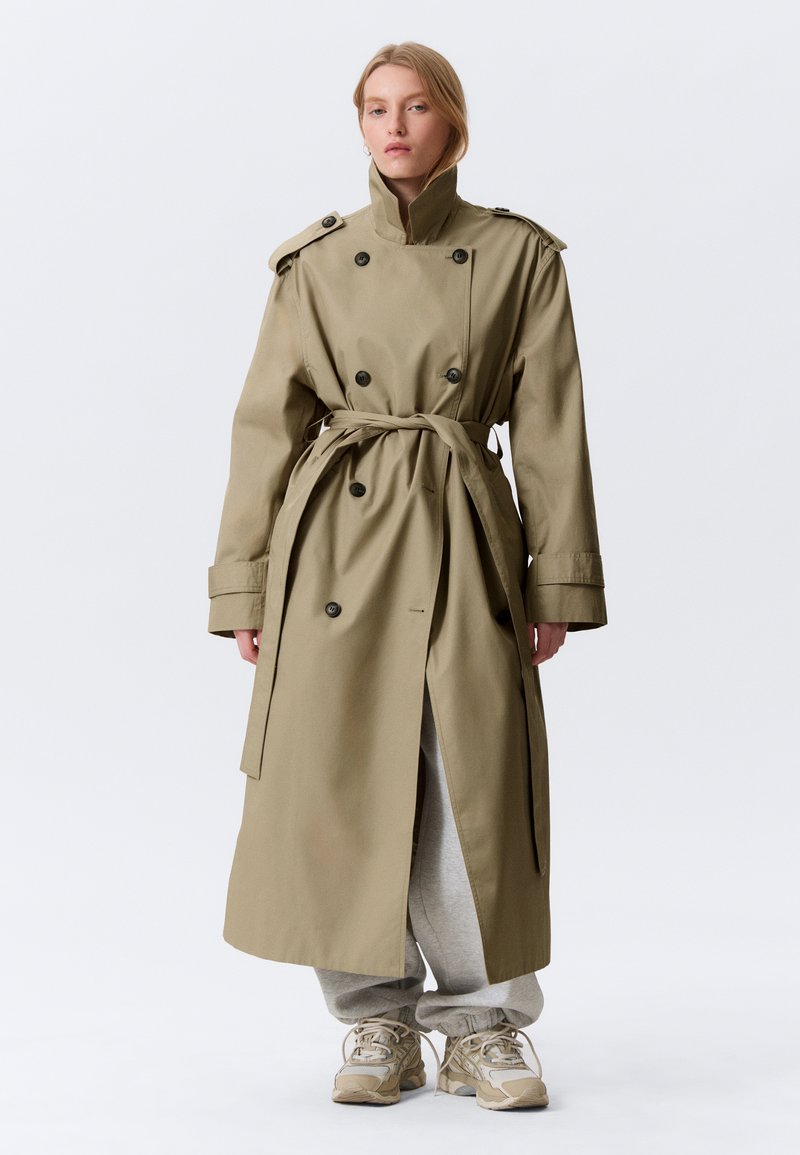 Femme debout portant un trench-coat beige ceinturé, un pantalon de survêtement gris et des baskets beiges, sur un fond clair uni.