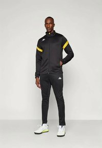 Μαύρο αθλητικό κοστούμι με κίτρινες λεπτομέρειες, περιλαμβάνοντας ζακέτα με φερμουάρ και εφαρμοστά παντελόνια. Κατασκευασμένο από λείο ύφασμα, με το λογότυπο Kappa στο στήθος και στα παντελόνια.