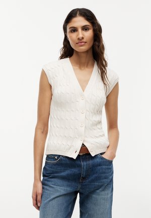 Femme portant un gilet sans manches blanc en maille torsadée à boutons et un jean bleu avec une ceinture marron, debout avec une main dans la poche.